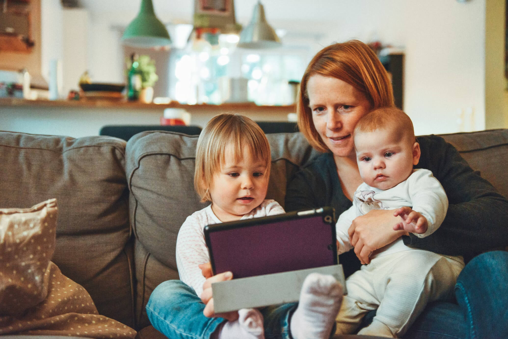 Woman, a toddler and a baby sitting on a sofa looking at a tablet together.