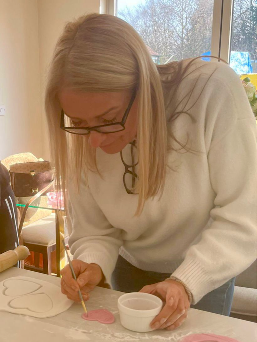 Image shows a woman with straight blond hair and dark rimmed glasses perched on her nose, another pair tucked into the top of her white winter jumper. She is bent over a table with a paint brush in one hand and a pot of paint in the other, decorating edible icing love heart shapes