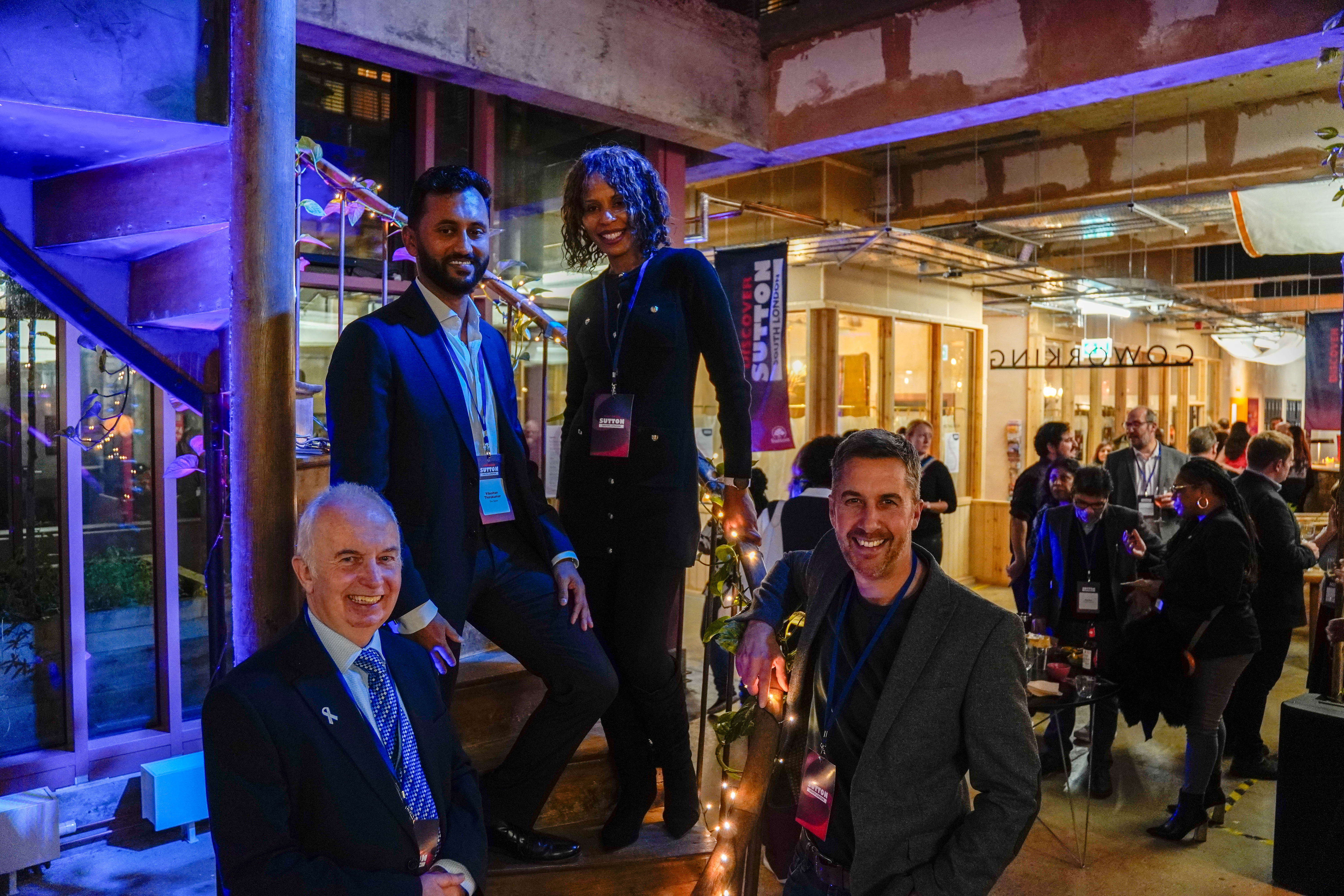 Leader of the Council, Councillor Barry Lewis at the launch of Sutton Councilu2019s Economic Growth Plan, standing with people from the event on a staircase with people talking in the background in a busy room.