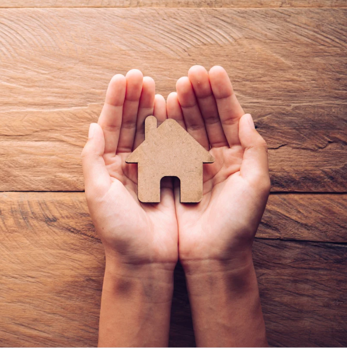 Image of horizontal wooden boards, with two arms reaching into the photo with outstretched hands holding a paper house 