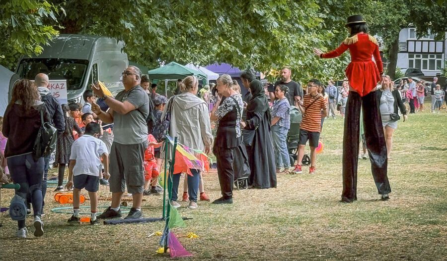 A gathering of people of different ages visiting community stalls on grass. To the right of the image there is a person on stilts in a red jacket.